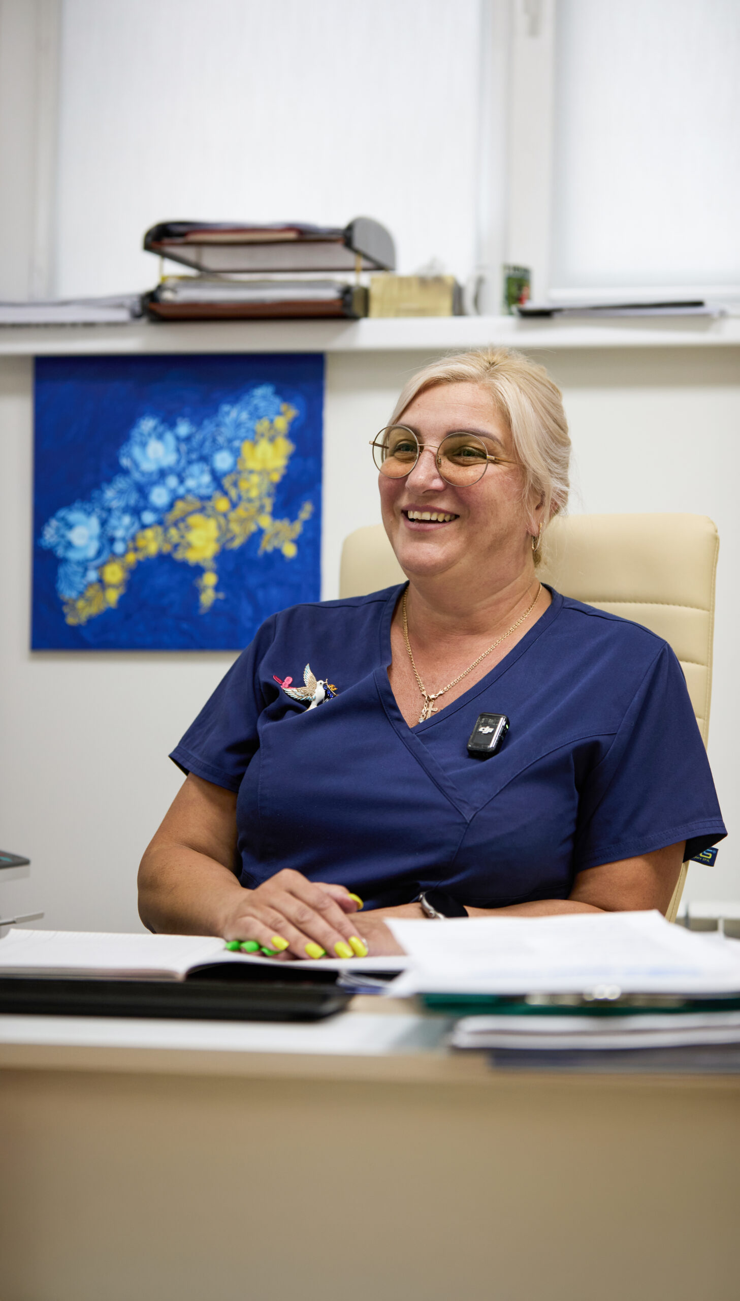 Жінка зі світлим волоссям, зібраним у низький хвіст, у круглих окулярах і темно-синьому медичному одязі. Катерина сидить у кріслі в кабінеті, на її обличчі — широка усмішка.