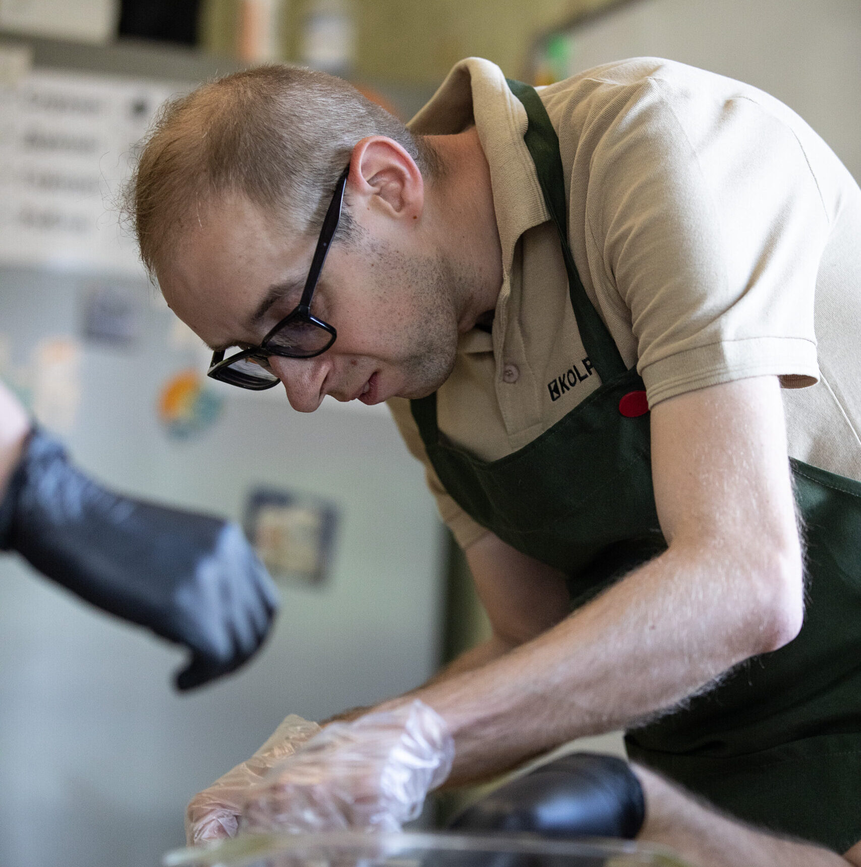 Чоловік із коротким світло-русявим волоссям, у темних окулярах і бежевій сорочці. Олександр працює над приготуванням страви за столом, нахилившись над робочою поверхнею.
