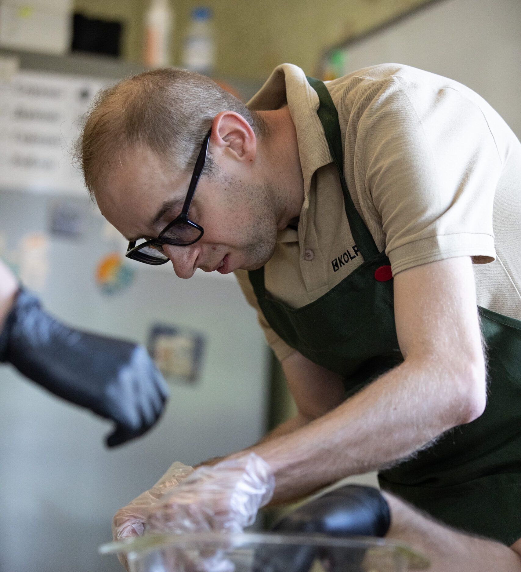 A man with short light brown hair, wearing dark glasses and a beige shirt. Oleksandr is preparing a dish at a table, leaning over the work surface.