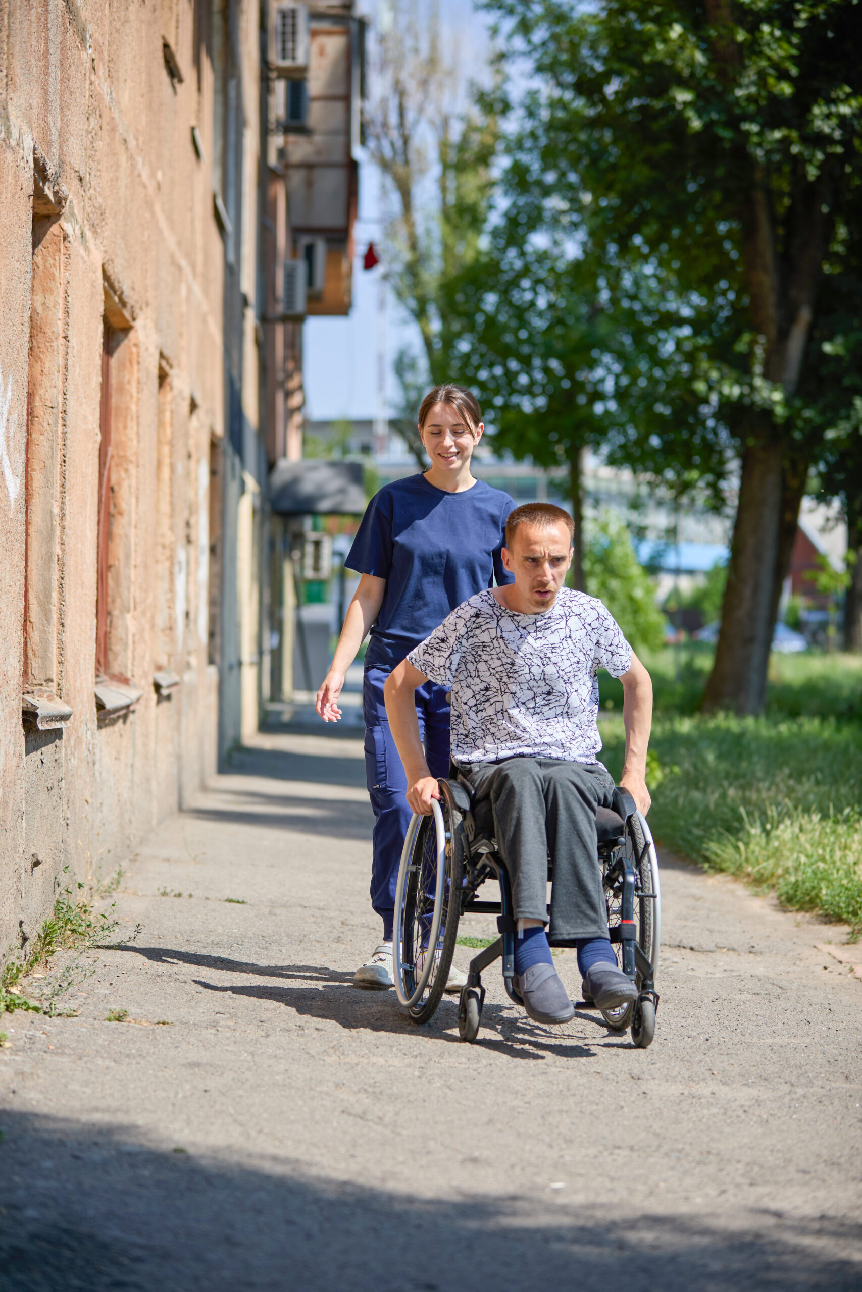 A young woman with brown hair tied back in a ponytail, wearing dark blue medical scrubs. A man using a wheelchair is beside Olha as they walk together through the grounds of a healthcare facility.