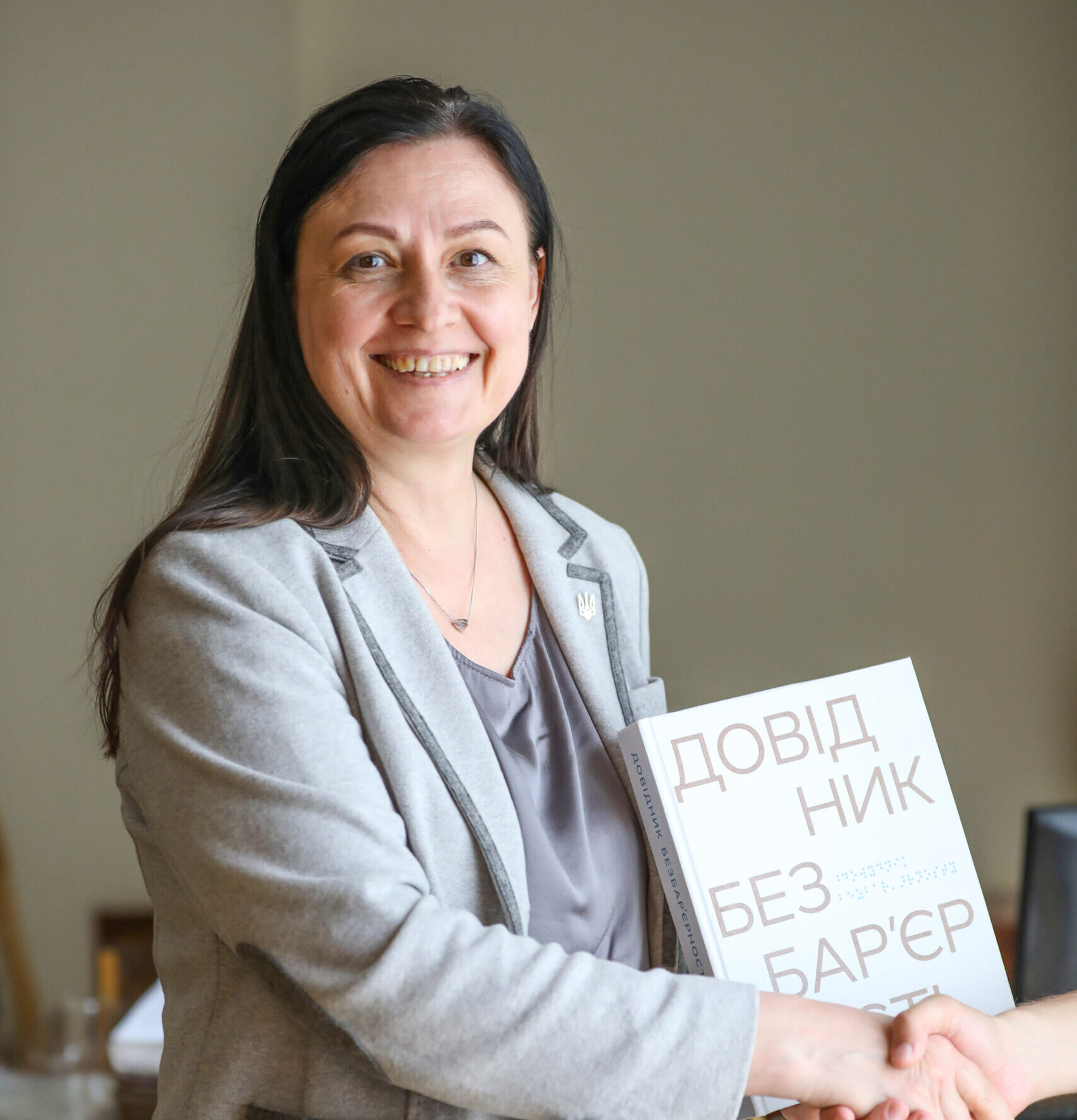 Жінка з довгим темним волоссям, одягнена у світло-сірий піджак і сіру блузку з V-подібним вирізом. Тетяна усміхається, тисне руку людині за кадром, а в іншій руці тримає довідник безбарʼєрності.