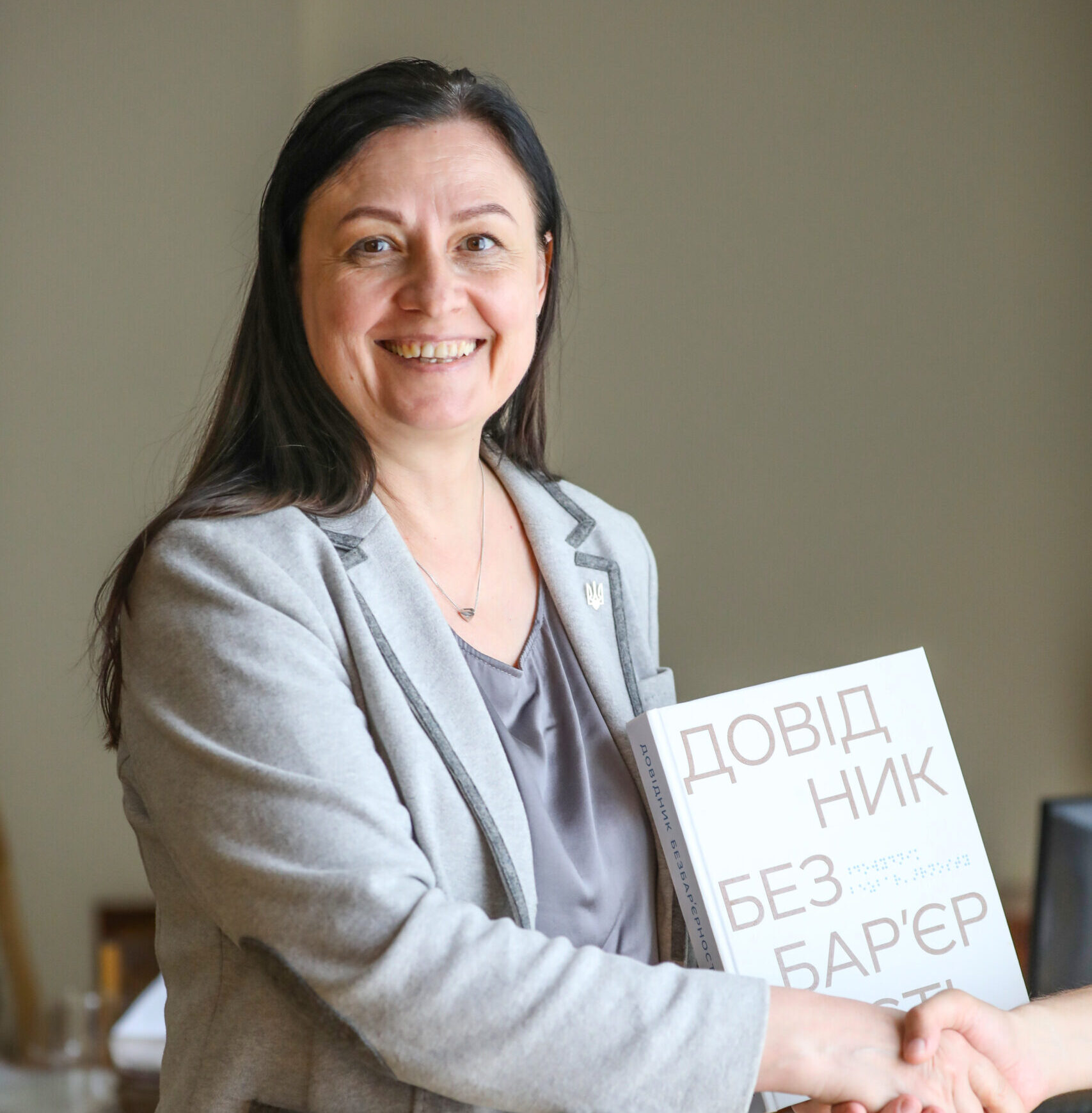 A woman with long dark hair, wearing a light grey blazer and a grey V-neck blouse. Tetiana is smiling as she shakes hands with someone off camera, holding an accessibility guidebook in her other hand.