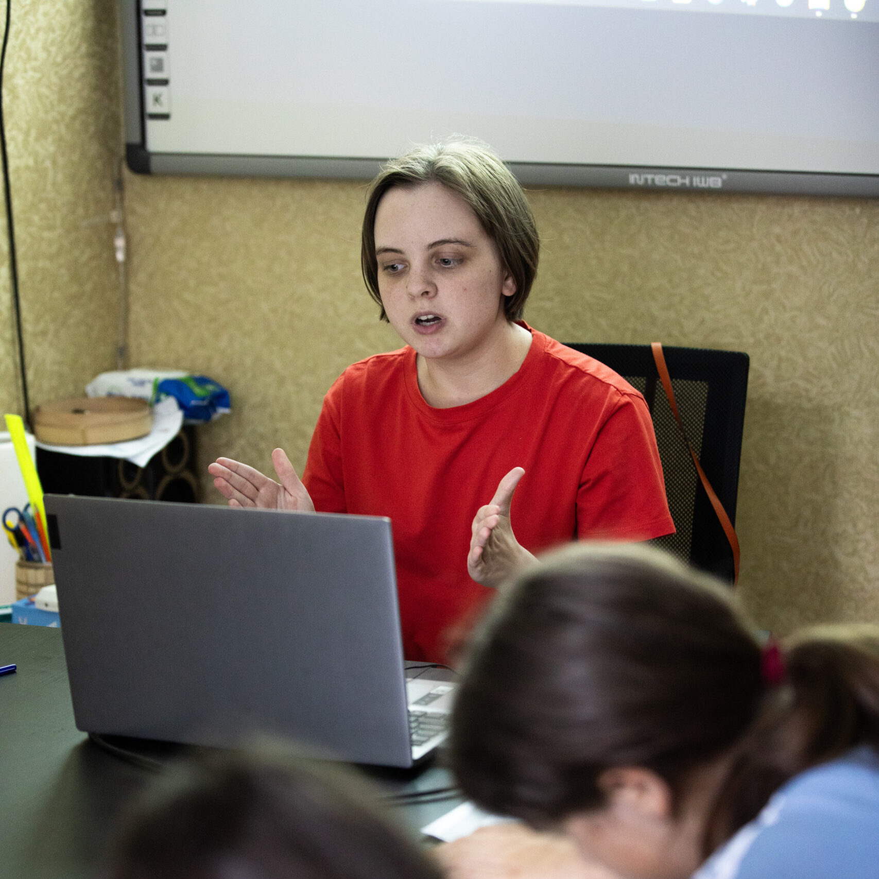 Молода жінка зі світло-русявим волоссям до плечей, одягнена в однотонну червону футболку. Тетяна сидить перед ноутбуком і активно жестикулює під час уроку; за її спиною видно навчальну смартдошку.