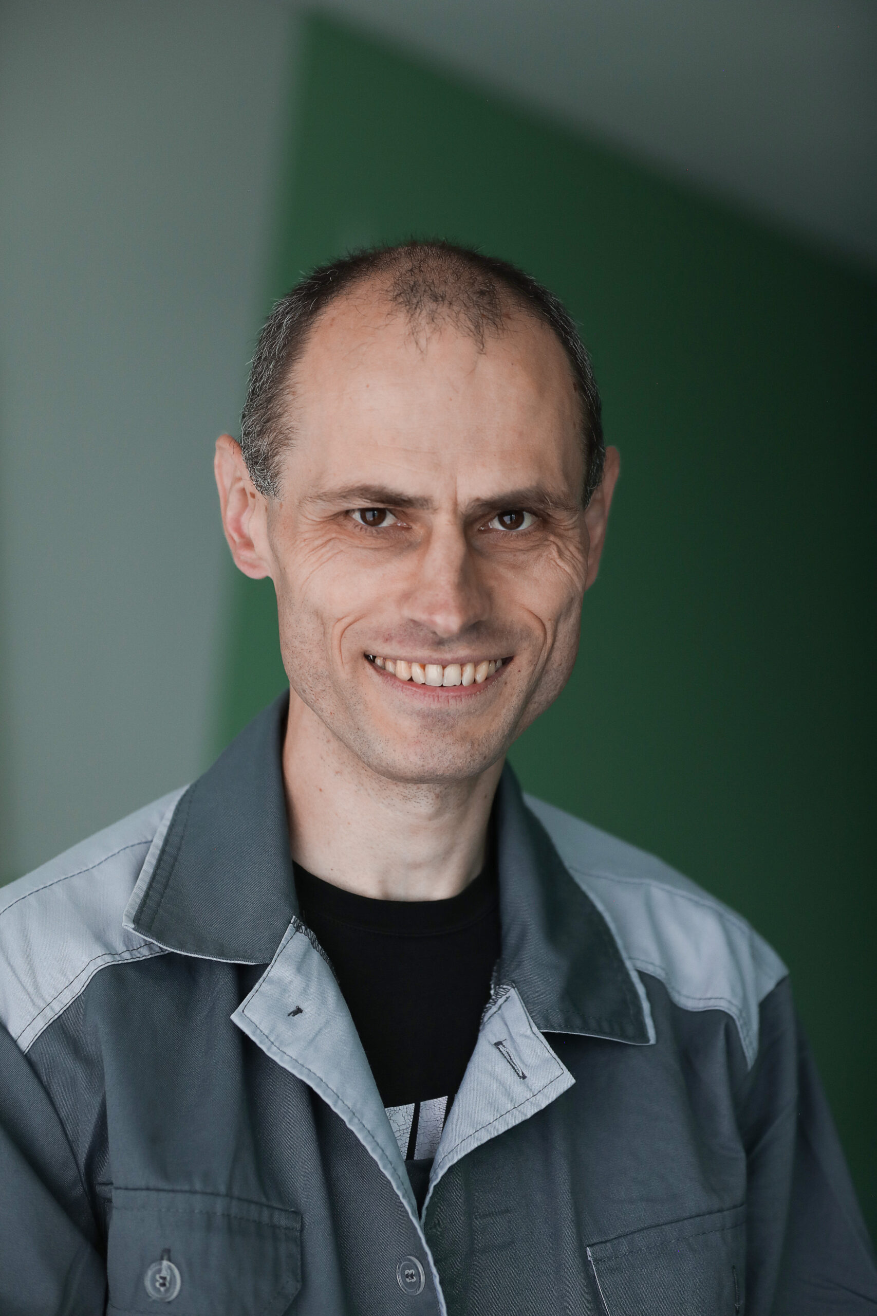 A man with short dark hair and no beard, with fair skin and brown eyes, wearing a grey work jacket over a black T-shirt. Vadym is standing in a back room and smiling at the camera.